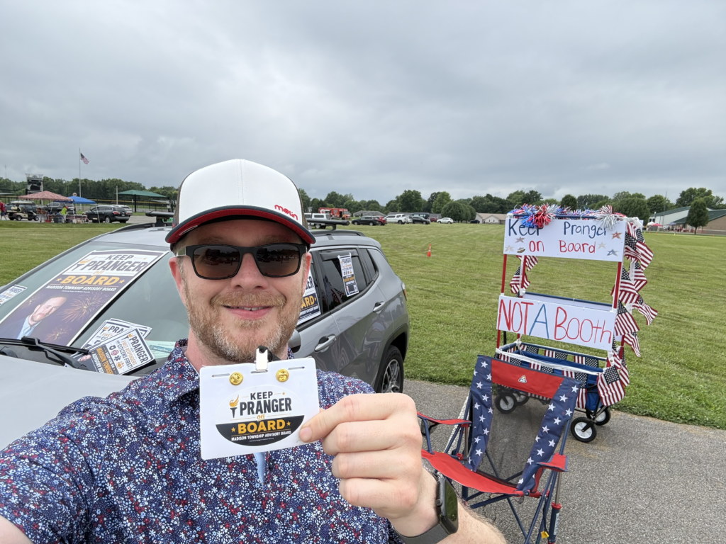 Candidate Pranger in sunglasses and a white cap takes a selfie while holding a “Keep Pranger on Board” campaign badge. Behind him, a gray SUV is covered with campaign signs, and a patriotic red, white, and blue booth decorated with American flags displays “Keep Pranger on Board” and “Not a Booth” signs at an outdoor community event.