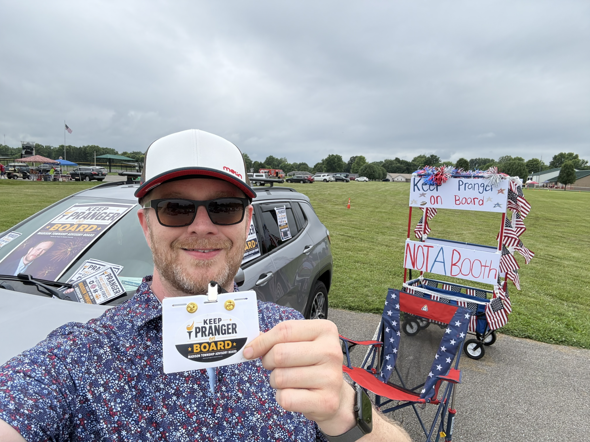 Candidate Pranger in sunglasses and a white cap takes a selfie while holding a “Keep Pranger on Board” campaign badge. Behind him, a gray SUV is covered with campaign signs, and a patriotic red, white, and blue booth decorated with American flags displays “Keep Pranger on Board” and “Not a Booth” signs at an outdoor community event.